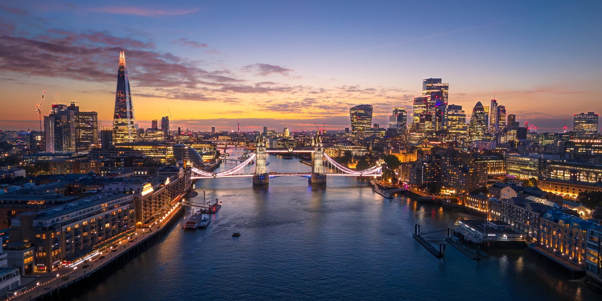 A stunning aerial panoramic view of London at twilight, featuring the Tower Bridge illuminated over the River Thames and the modern City skyline glowing in the background