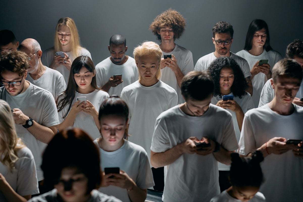 Group of diverse people wearing white t-shirts, looking down at mobile devices against a gray background.